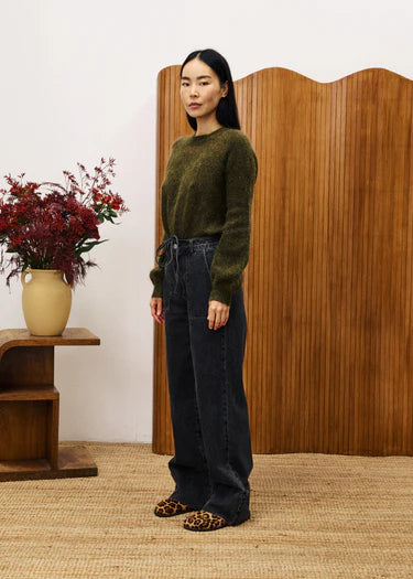 Woman wearing a green sweater and dark jeans standing in a room with wooden furniture and flowers.