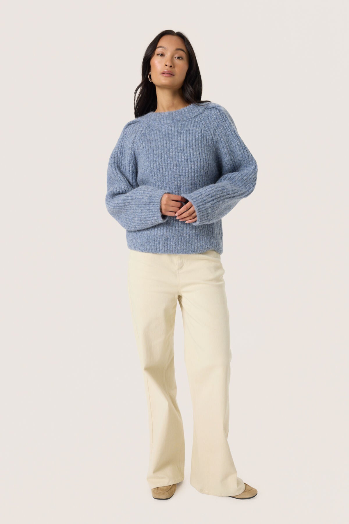 Woman wearing a blue sweater and white pants on a light gray background