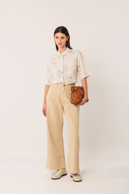 Woman wearing a beige blouse and pants holding a brown leather clutch against a white background