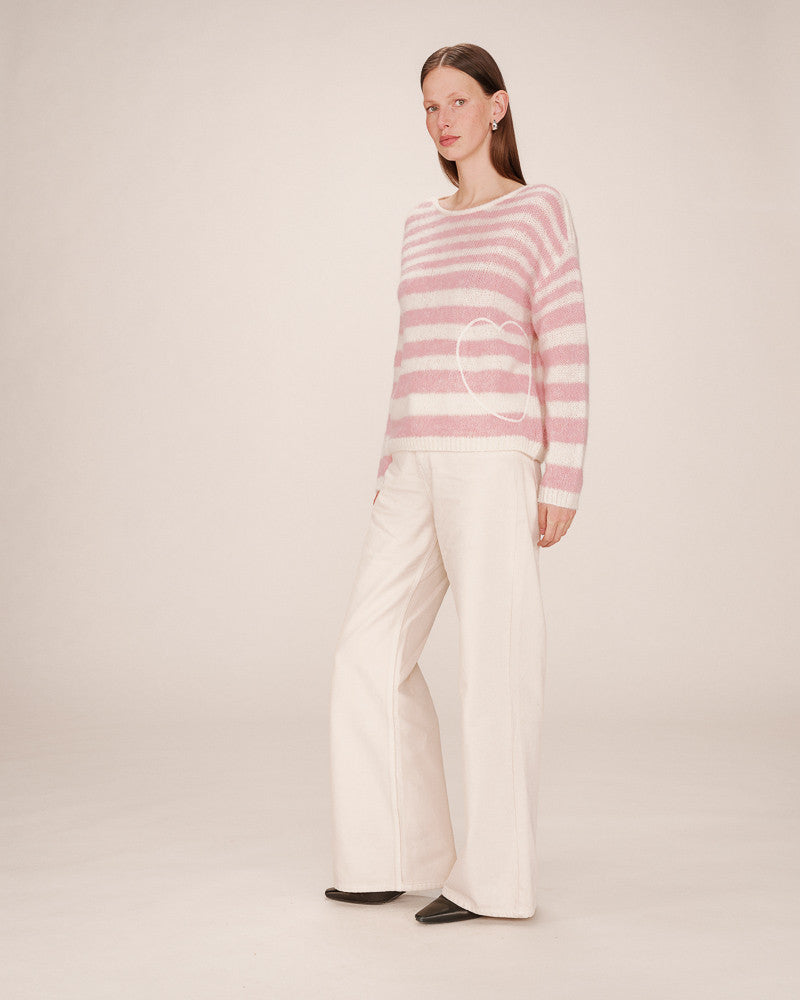 Woman wearing a pink and white striped sweater with a heart design, paired with white pants on a plain background.