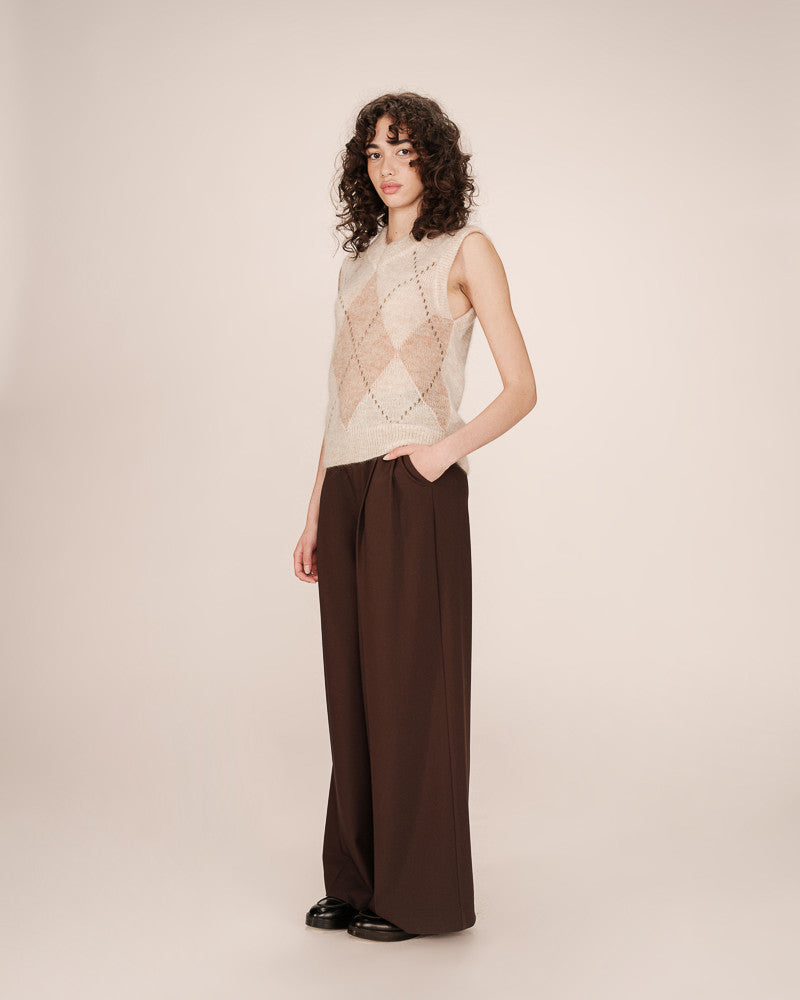 Woman wearing a sleeveless patterned top and wide-leg brown pants on a plain background