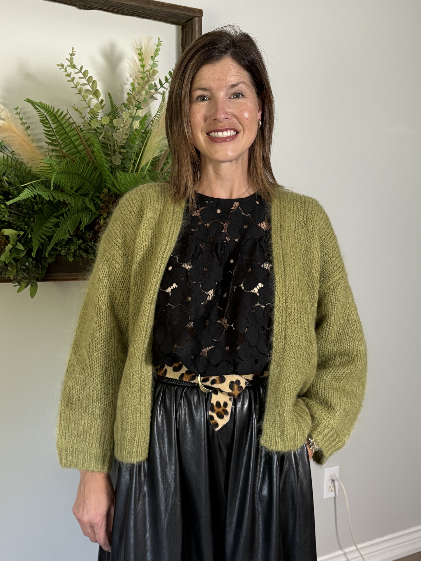 Woman wearing a green cardigan over a black top with animal print, standing in front of a plant.