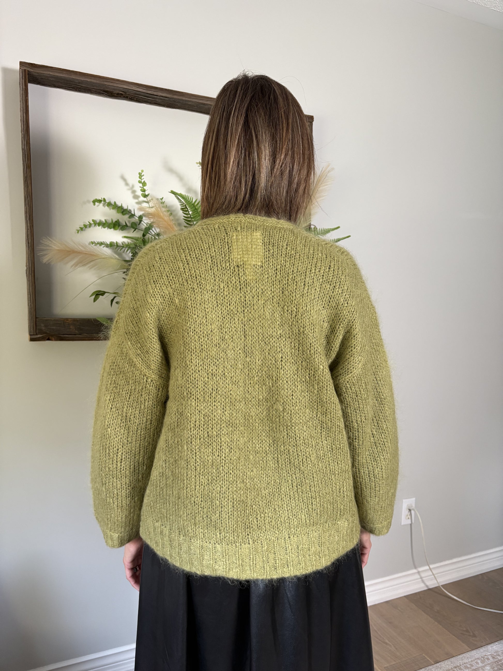 Person wearing a green knitted sweater standing in front of a wall with a framed picture.