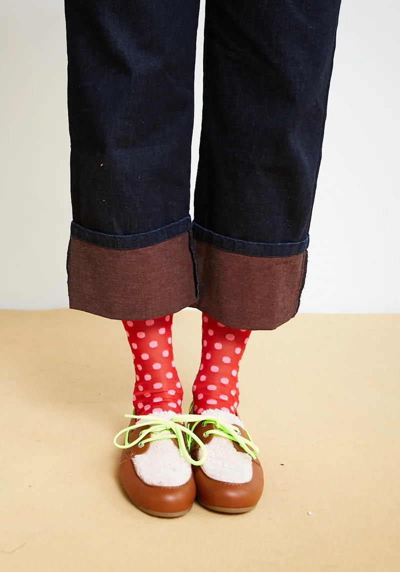 A person's legs wearing dark blue denim jeans with rolled cuffs showing brown lining, red polka-dot socks, and brown leather moccasin-style shoes with white fur trim and neon green laces, standing against a plain beige wall and floor.