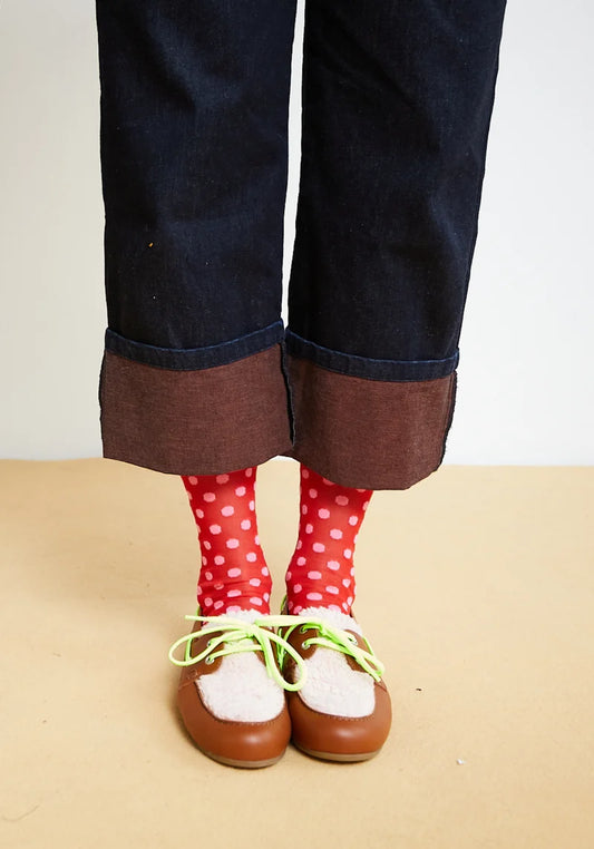A person's legs wearing dark blue denim jeans with rolled cuffs showing brown lining, red polka-dot socks, and brown leather moccasin-style shoes with white fur trim and neon green laces, standing against a plain beige wall and floor.