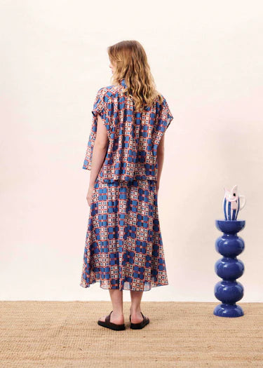 Woman wearing a patterned dress standing next to a blue vase on a wooden floor.