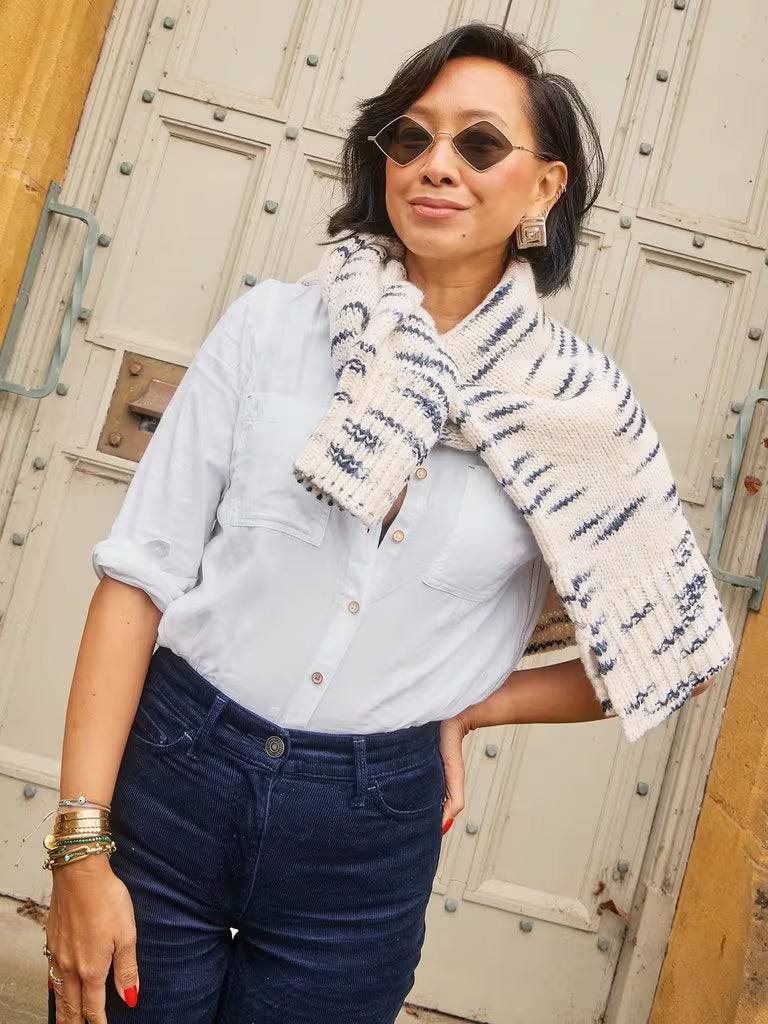 Woman wearing a light blue shirt, dark jeans, and a patterned scarf in front of a textured wall.