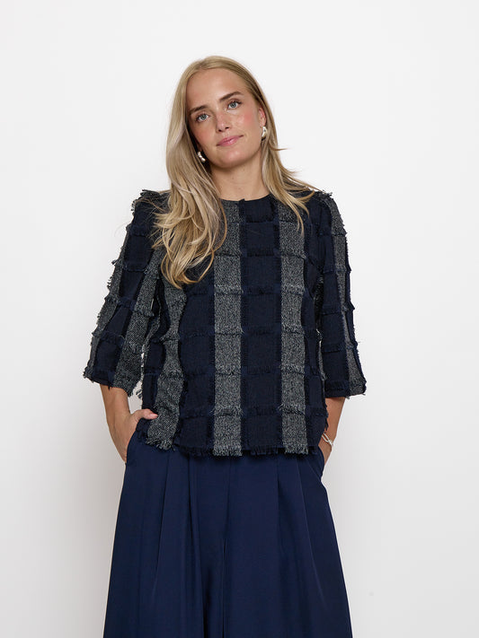 A woman modeling a textured, patchwork-style navy blue and gray cropped jacket with ruffled sleeves, paired with a navy blue skirt, against a plain white studio background.
