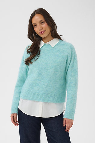 A woman wearing a light mint green knit sweater with a white pointed collar, layered over a white collared shirt and dark blue jeans, against a plain light gray studio background.