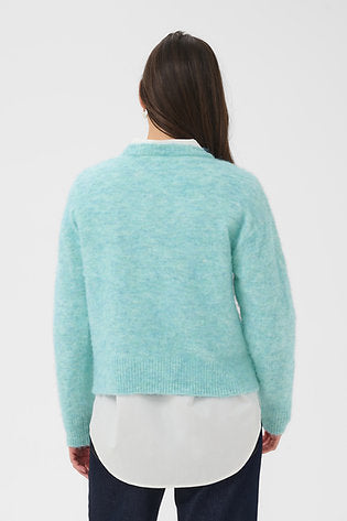 A woman viewed from the back wearing a light mint green knit cardigan over a white collared shirt and dark navy pants, against a plain light gray studio background.