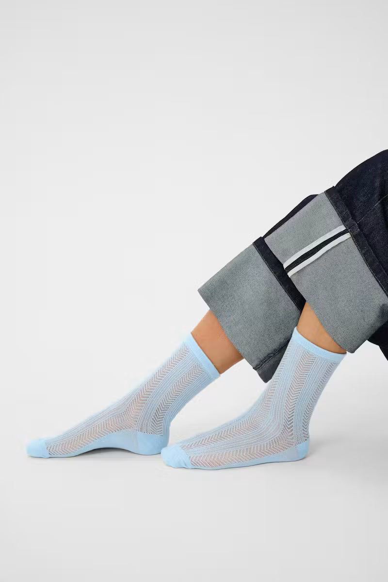 A pair of light blue ankle socks with a textured knit pattern, worn on a person's feet with dark gray and navy cuffed jeans, against a light gray studio background.