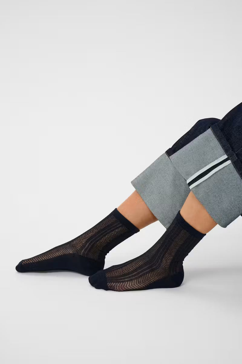 A close-up of a person's legs wearing black sheer ribbed ankle socks with black toe caps, paired with gray and navy cropped trousers, against a light gray studio background.