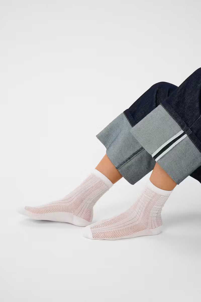 A close-up of a person's feet wearing white ribbed knit ankle socks with dark cuffed jeans, photographed against a clean light gray studio background.