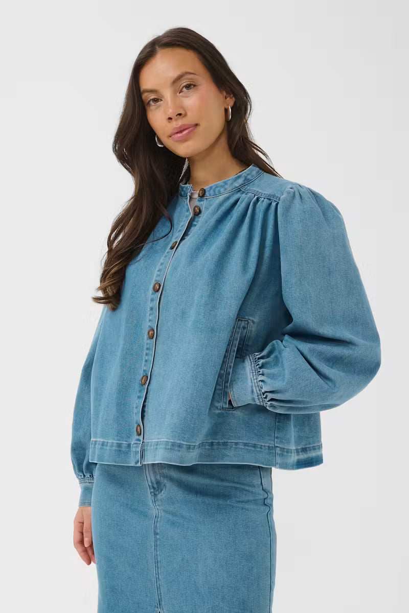 A woman modeling a cropped, light blue denim jacket with voluminous long sleeves, a high gathered neckline, and brown buttons, paired with a matching denim skirt against a light gray studio background.