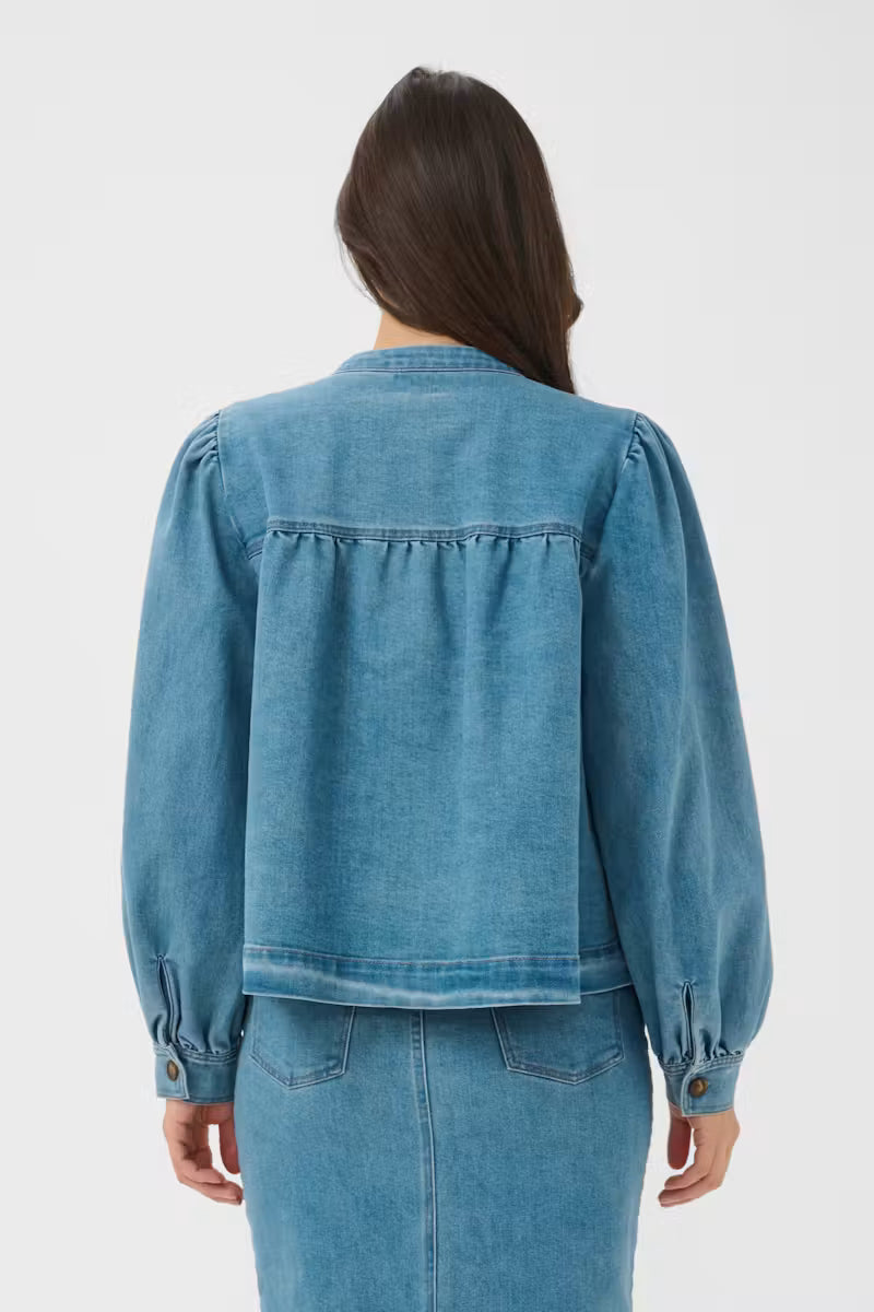 A woman viewed from the back wearing a cropped blue denim jacket with gathered puff sleeves and a matching denim skirt against a light gray studio background.