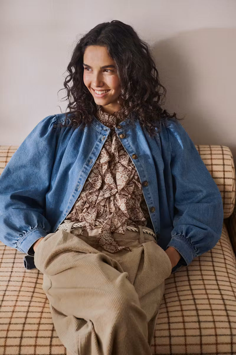 A woman with long, curly dark hair sits on a beige plaid couch, wearing a blue denim jacket over a brown patterned blouse and beige corduroy trousers.