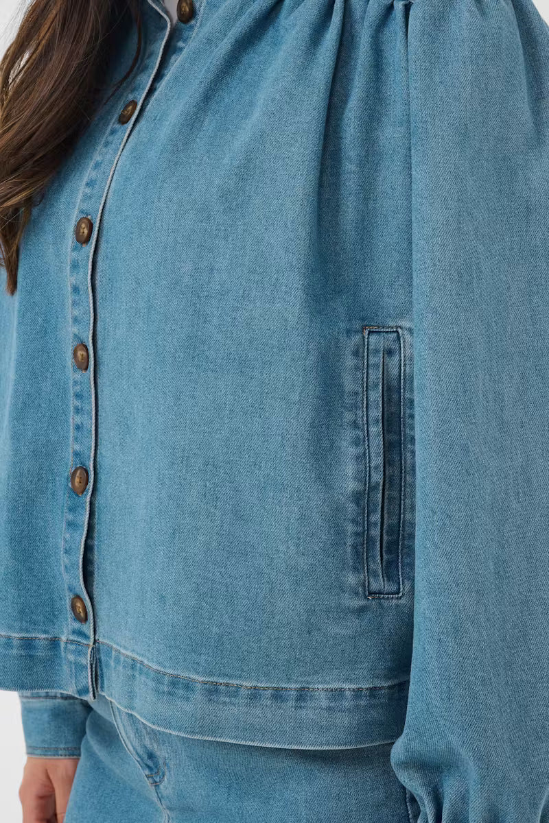 Close-up of a person wearing a light blue denim jacket with a vertical chest pocket and brown buttons, paired with matching blue denim bottoms.
