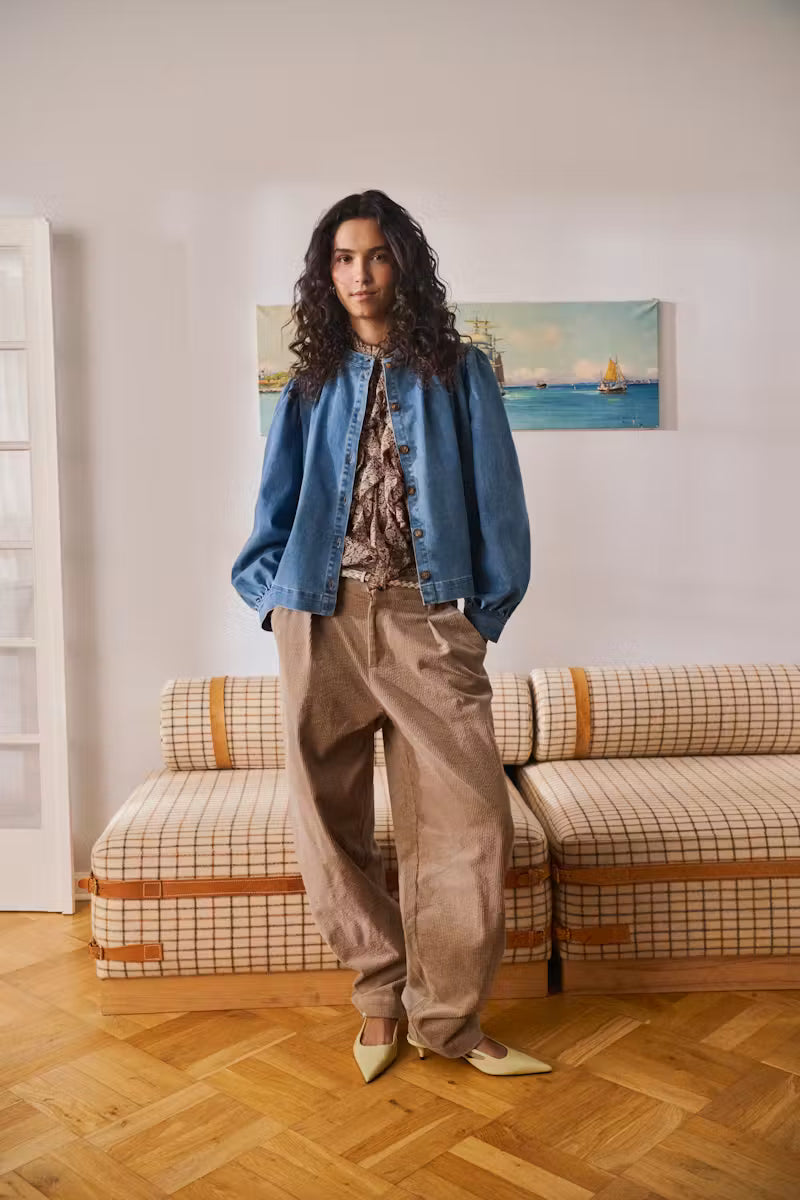 A model stands in a room wearing a blue denim jacket, a patterned blouse, loose brown corduroy trousers, and cream pointed-toe flats. Behind her is a plaid sofa and a framed painting on a light wall.