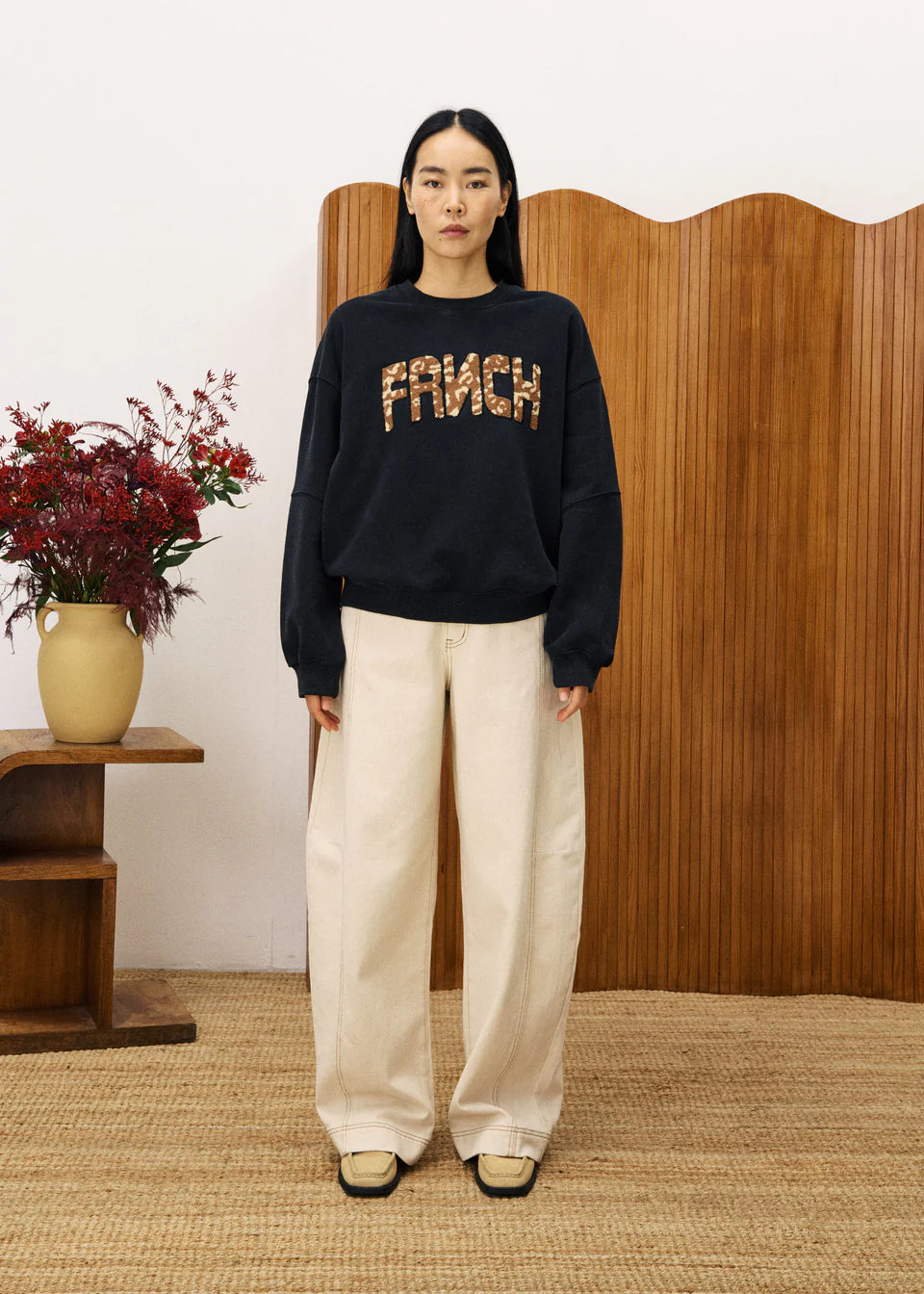 Person wearing a black sweater with 'FRAICH' text, standing in a room with wooden paneling and a vase of flowers.