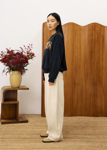 Woman wearing a dark sweater and light pants standing in a room with a wooden screen and vase of flowers.