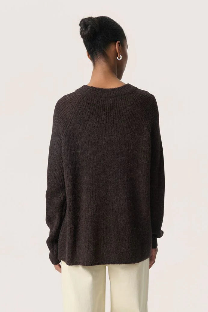 Person wearing a dark brown sweater and white pants on a plain background