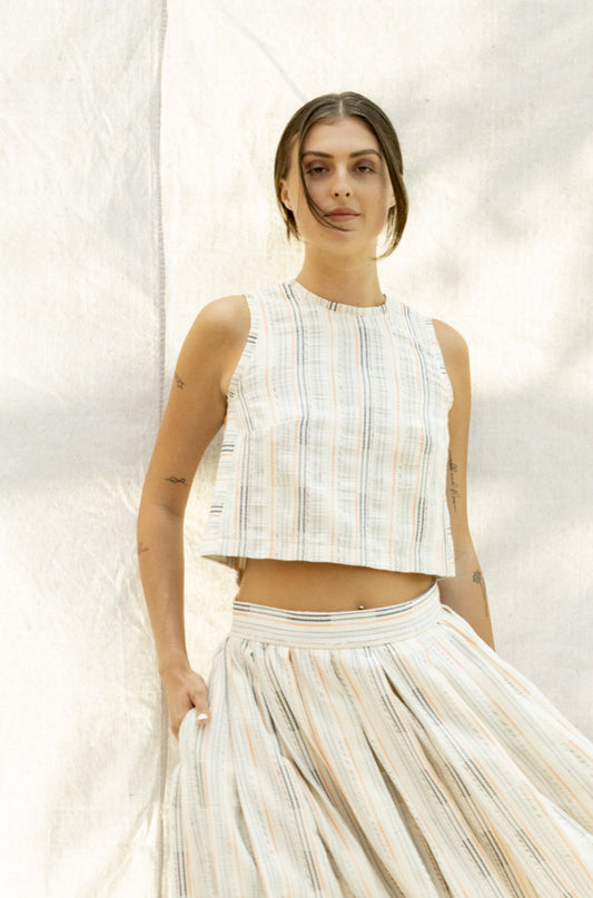 Tulum Top, a tailored fit crop top with a playful sleeveless design and colourful accents. Featuring a crew neck adorned with a beautiful zipper detail, this top is made from a unique fabric that combines blue and orange stripes with metallic blue and pink accents on a cream background. Its loose construction adds texture and allows for breathability, making this top perfect for any fun and chic outfit.
