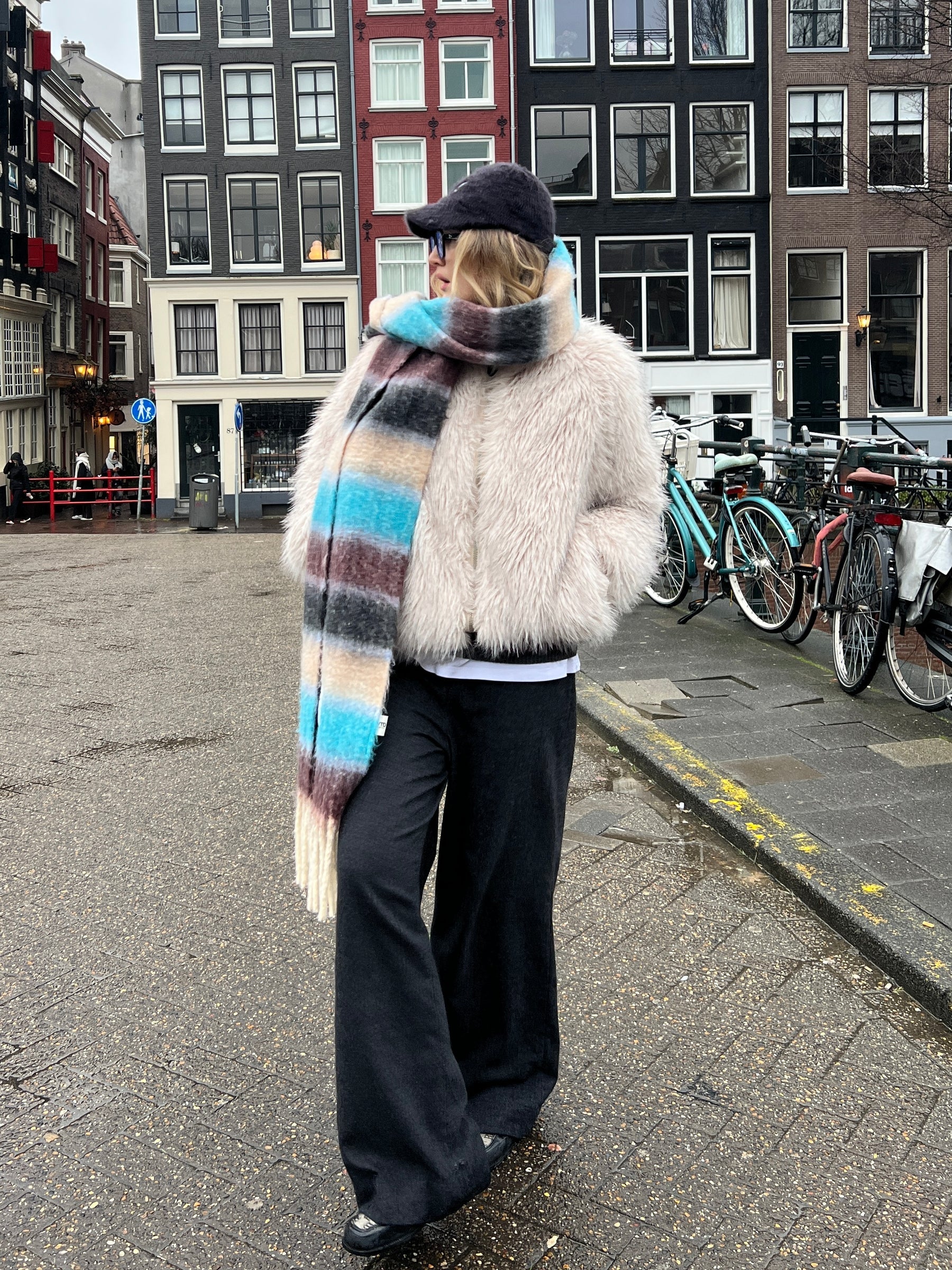 Person wearing a colorful scarf and fur coat on a street with bicycles and buildings in the background