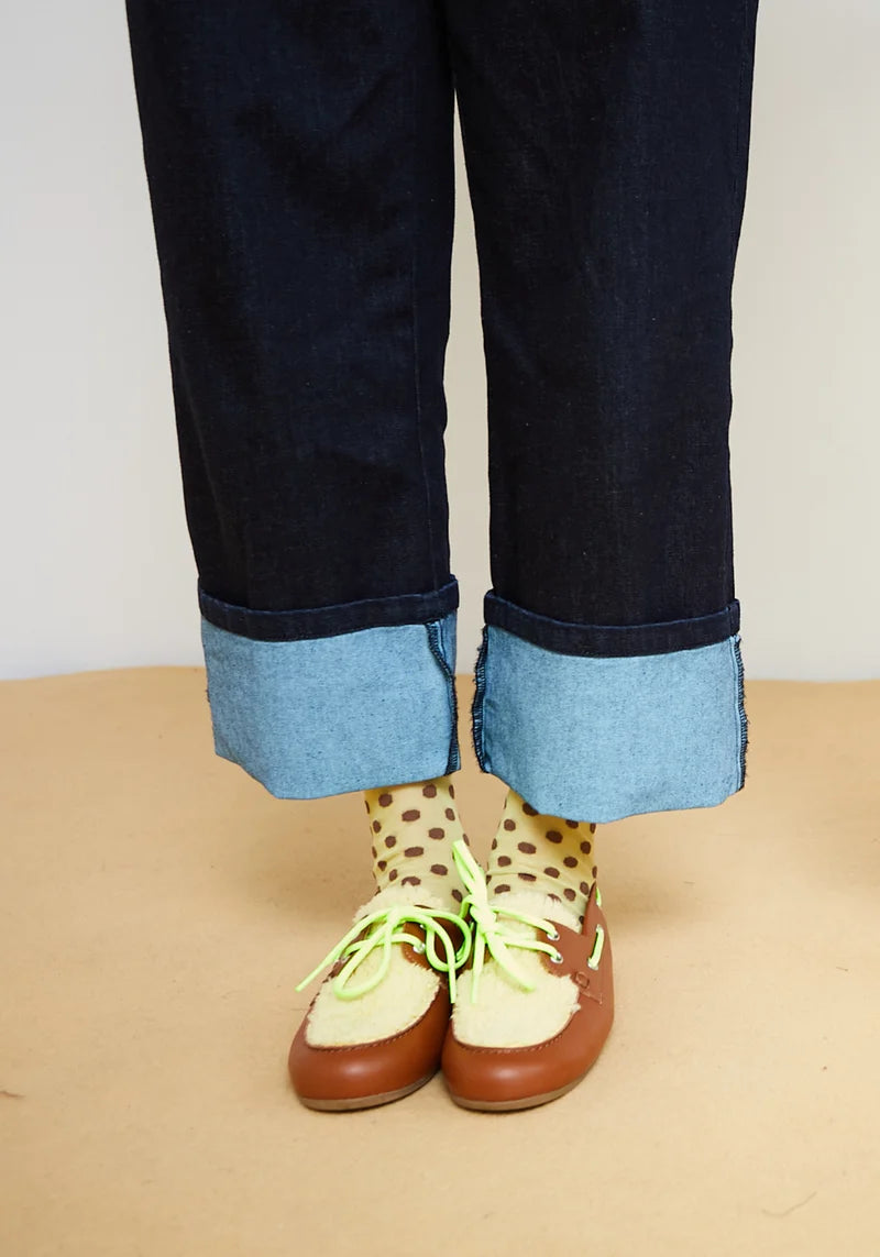 A close-up of a person's legs from the knees down, wearing dark blue cuffed jeans, beige socks with brown polka dots, and brown shoes with bright green laces, standing on a light tan floor against a white wall.